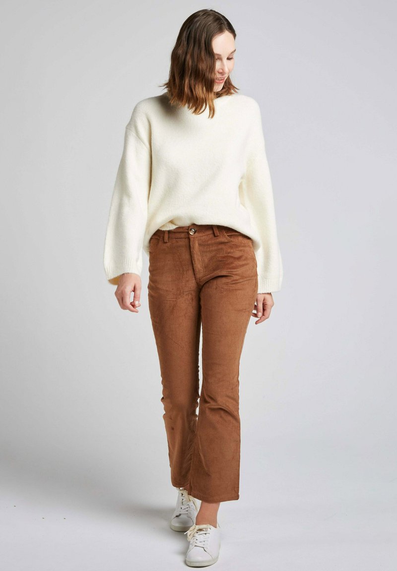 Pull blanc texturé avec des manches ballon, associé à un pantalon en velours côtelé marron à pattes d'éléphant et des baskets blanches. Taille ajustée avec un détail de bouton.