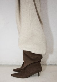 Stivaletti alla caviglia in camoscio marrone, morbidi, con punta appuntita e tacchi kitten, indossati con pantaloni beige a gamba larga su sfondo di parete chiara.