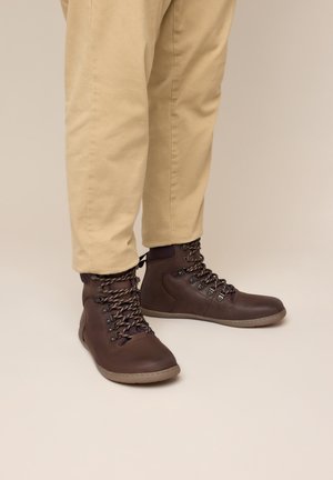 Person, die beige Hose und dunkelbraune knöchelhohe Lederstiefel mit gemusterten Schnürsenkeln trägt, steht vor neutralem Hintergrund.