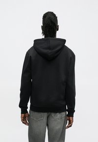 Sweatshirt preta com capuz em mistura de algodão, com cordão no capuz, punhos e barra canelados. Usada com jeans cinzentos, vista por trás.