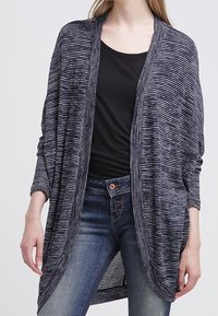 Cardigan leggero a righe navy con apertura frontale, maniche lunghe e design drappeggiato abbinato a un top nero e jeans blu.