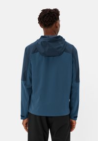 Giacca leggera da uomo di colore blu scuro, con cappuccio, spalle senza cuciture e maniche di un colore più scuro in contrasto. Texture liscia e design sportivo.