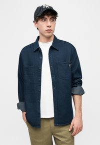 Νεαρός άνδρας με λευκό μπλουζάκι, σκούρο μπλε πουκάμισο με κουμπιά και μανίκια γυρισμένα, χακί παντελόνι και μαύρο καπέλο του μπέιζμπολ με ρίγες, κοιτάζει πλάγια.