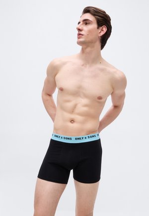 Νεαρός άνδρας με σκοτεινά μαλλιά που φοράει μαύρα ανδρικά εσώρουχα boxer με ανοιχτά μπλε ζώνη μέσης με την ετικέτα "ONLY & SONS", στέκεται μπροστά σε απλό φωτεινό φόντο.