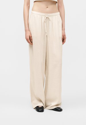 Frau trägt lockere, hoch taillierte beige Weitbein-Hosen mit Kordelzug und schwarzen flachen Schuhen, steht vor einem schlichten Hintergrund.