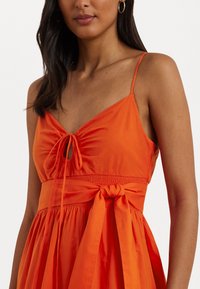 Vestido laranja com decote amarrado e recorte em forma de olho. Apresenta uma cintura franzida e uma saia fluida. Alças finas. Tecido suave.