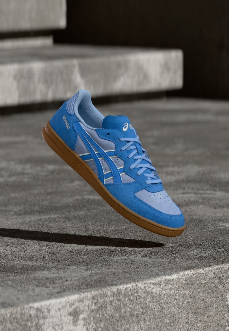 Zapatilla deportiva azul que combina materiales de ante y malla, con detalles perforados, diseño de cordones y suela de goma.