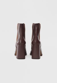 Bottines en cuir marron avec un talon carré haut. L'arrière présente une finition lisse et des panneaux latéraux contrastés en tan pour ajouter de la texture.