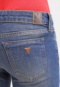 Siniset farkut, joissa on ruskea nahkapatchi, kolmiomainen logo takataskussa ja näkyviä ompeluyksityiskohtia.