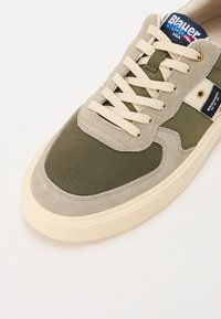 Grønne og beige sneakers laget av semsket skinn og mesh-materialer, med kremfargede snører, en teksturert såle og et logolapp på tungen.