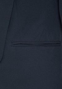 Marinblå kavajtyg med en slät textur, subtil töjbarhet, samt en enda, horisontell ficka på vänster sida.