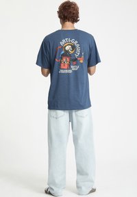 Camiseta de algodón azul marino con un estampado gráfico en la parte posterior, que incluye el texto "ANTI-GRAVITY BOTTLE SHOP" y una imagen colorida. Corte relajado.