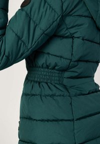 Giubbotto imbottito di colore verde scuro, con texture trapuntata, vita elastica e cappuccio. Include una tasca laterale con dettagli in zip.