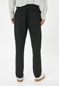 Sorte bukser med elastisk talje og en glat overflade, der har to baglommer og en tapered ben-design. Båret med hvide sneakers.