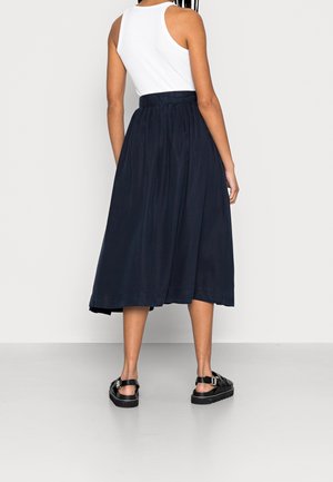 Vrouw draagt een witte mouwloze top, een marineblauwe midirok en zwarte sandalen met gespen, staande tegen een effen lichte achtergrond.