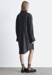 Schwarzes und weiß gestreiftes Hemdkleid mit langen Ärmeln, Knopfmanschetten, einem elastischen Bund und einem leicht längeren Rückensaum. Getragen mit schwarzen Schuhen und braunen Socken.