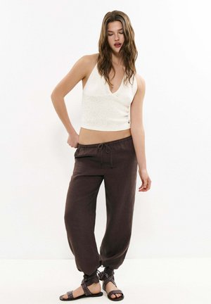 Junge Frau mit langen Haaren, die ein weißes ärmelloses Stricktop, dunkle lockere Hose und schwarze Sandalen mit Riemchen trägt, steht vor einem schlichten Hintergrund.