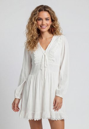 Donna sorridente con capelli mossi che indossa un vestito bianco a maniche lunghe con dettagli in pizzo e orecchini floreali marroni su sfondo semplice.