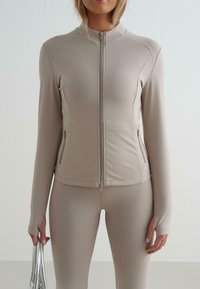 Beige Zip-Jacke mit langen Ärmeln und tailliertem Schnitt. Mit seitlichen Taschen und dehnbarem Stoff, kombiniert mit passenden Leggings.