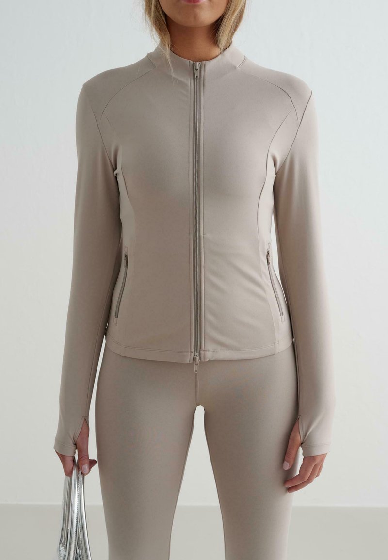 Beige Zip-Jacke mit langen Ärmeln und tailliertem Schnitt. Mit seitlichen Taschen und dehnbarem Stoff, kombiniert mit passenden Leggings.