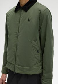Giacca bomber verde con colletto in corduroy nero, chiusura a zip e una tasca frontale con dettaglio bottone. Design semplice e texture liscia.