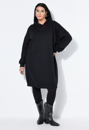 Vrouw met lang donker haar die een losse zwarte capuchonjurk, zwarte legging en zwarte gestudeerde cowboylaarzen draagt, staande tegen een witte muur.