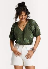 Haut vert à manches courtes avec une texture à pois et une broderie florale colorée ; associé à un short blanc taille haute avec des coutures bleues.