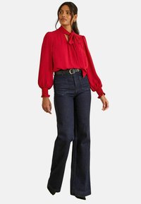 Piros blúz hosszú ujjal és nyakkendős részlettel, párosítva magas derekú sötét denimből készült cseppnadrággal és fekete övvel.