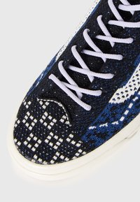 Sportsko sko med et blått, svart og hvitt mønstrete tekstiloverdel, med hvite lisser og en strukturert tåhette med avrundet design.