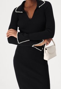 Svart ribbad klänning med V-ringning samt krage och manschetter accentuerade med pärlor. Har en figurnära silhuett. Håller en liten handväska i krämfärg.