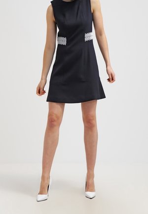 Ärmelloses schwarzes A-Linien-Kleid mit Perlenverzierungen an der Taille, getragen mit weißen spitzen High Heels auf neutralem Hintergrund.