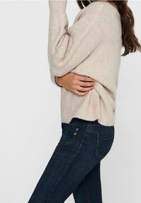 Pull beige tricoté à coupe ample et texture côtelée, associé à un jean skinny bleu foncé avec poches avant et arrière.