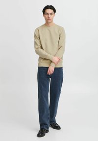 Beige textuurtrui met lange mouwen, gecombineerd met donkere blauwe wijde jeans en zwarte schoenen. Het model staat rechtop met de armen gekruist.