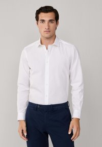 Camisa blanca de manga larga con cuello abotonado. Presenta un corte ajustado y una textura suave. Combinada con pantalones oscuros en color marino.