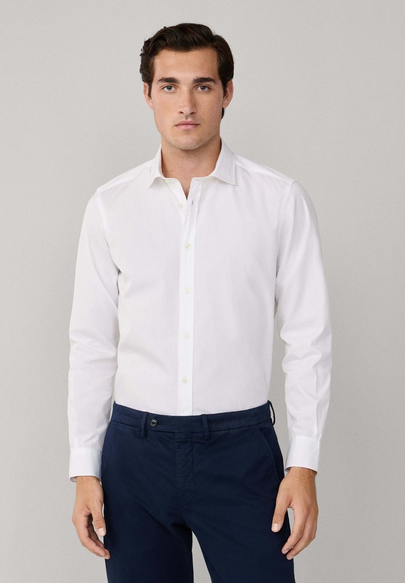 Camisa blanca de manga larga con cuello abotonado. Presenta un corte ajustado y una textura suave. Combinada con pantalones oscuros en color marino.