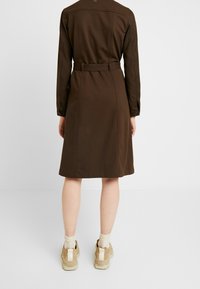 Ruskea, polvipituinen mekko, jossa on pitkät hihat ja vyöllä varustettu vyötärö; valmistettu sileästä kankaasta yksinkertaisella suunnittelulla. Yhdistetty beigeihin tennareihin ja sukkiin.