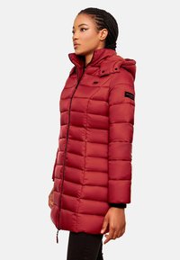 Red padded coat with a high collar and hood, featuring a zip closure, side pockets, and a logo detail on the sleeve. Smooth fabric texture.