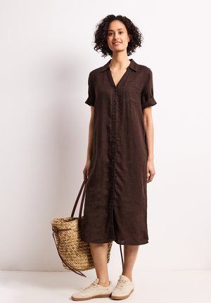Kobieta stojąca z kręconymi włosami, ubrana w brązową sukienkę midi na guziki, białe trampki i trzymająca plecioną słomianą torbę.