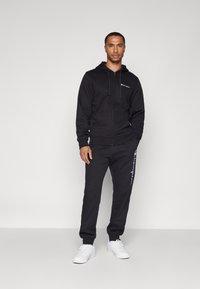 Sorte zip-up hettegenser og matchende joggebukser laget av bomull. Har logo på brystet og siden, med ribbestrikkede mansjetter og midjebånd. Hvite joggesko.