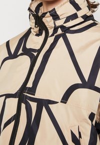 Giacca beige con motivi astratti neri, colletto alto e chiusura con zip. Presenta una texture liscia e tasche laterali. Design leggero.