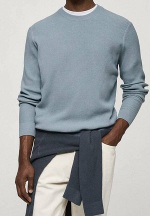 Mand iført lyseblå ribbet sweater over hvid skjorte, med mørkeblå sweater bundet om taljen og hvide bukser.