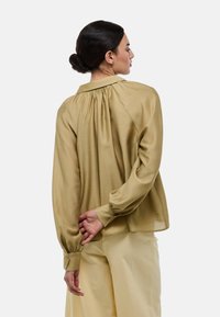 Blouse vert olive, ample, avec un dos froncé, des manches longues et des poignets boutonnés. Tissu léger avec une texture lisse.