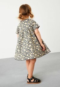 Graues Kleid mit Blumenmuster, einem ausgestellten Rock und kurzen Ärmeln. Kombiniert mit schwarzen Riemchensandalen. Detail mit gewelltem Saum.
