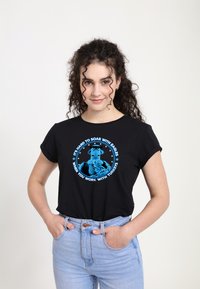 Černé tričko s krátkým rukávem s modrým grafickým designem a textem o orlech a krůtách. Model nosí světle modré vysoké džíny.