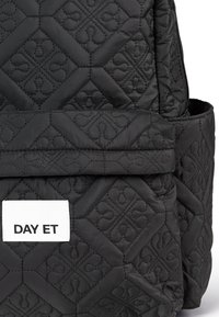 Sac à dos noir matelassé avec un motif géométrique, doté d'une poche zippée à l'avant et d'une étiquette blanche avec "DAY ET". La surface texturée met en valeur les détails.