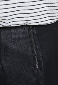 Svart läderkjol med en texturerad yta. En silvrig dragkedja löper vertikalt på ena sidan. Tunt tyg i marinblått och vitt med ränder ovanpå.