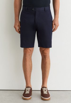 Muž v námorníckych modrých šortkách na mieru a hnedých semišových teniskách stojaci pred jednoduchou smotanovou stenou.