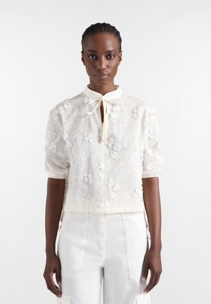 Kobieta ubrana w białą, kwiatowo haftowaną bluzkę z bufkami na rękawach i wiązanym kołnierzykiem, w zestawieniu z białymi spodniami z wysokim stanem.