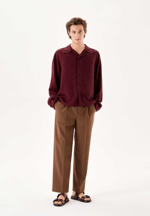 Chemise boutonnée bordeaux avec un col, associée à un pantalon plissé marron et des sandales foncées. Tissu lisse et coupe décontractée.
