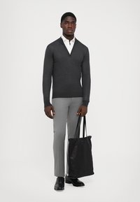 Grauer Zip-Hals-Pullover, schmal geschnittene graue Hose und schwarze Lederschuhe. Hält eine schwarze Tragetasche. Einfaches, modernes Design.